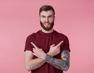 Wall Mural - Portrait of young handsome frowning misunderstanding red bearded man in red t-shirt, stands over pink background looks at the camera with disgust and points in different directions.