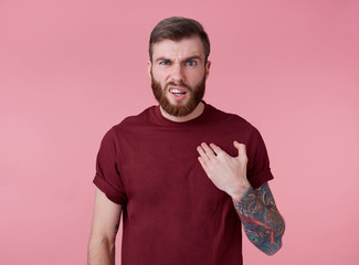 Wall Mural - Portrait of young handsome frowning misunderstanding red bearded man in red t-shirt, stands over pink background looks at the camera with disgust.