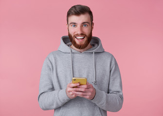 Wall Mural - Young handsome positive red bearded man in gray hoodie, looks happy and broadly smiles, holding smartphone, stands over pink background.