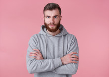 Photo Of Young Frowning Handsome Red Bearded Man In Gray Hoodie, Stands With Crossed Arms, Disapprovingly Looks At Camera Wit Raised Eyebrow ,stands Over Pink Background.