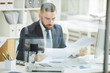 © Seventyfour - Serious young bearded businessman in suit sitting at table and examining financial papers in office