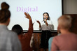 © Seventyfour - Portrait of contemporary businesswoman giving presentation standing by word DIGITAL on projector screen and talking to audience smiling cheerfully, copy space