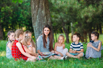 Children and education, young woman at work as educator reading book to boys and girls in park.