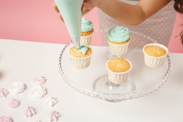 Wall Mural - Close up cropped housewife female chef cook confectioner or baker in apron white t-shirt decorating cake cupcake at table isolated on pink pastel background in studio. Mock up copy space food concept.