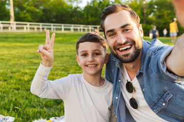 Young man having fun with his little brother or son outoors in park beautiful green grass take a selfie by camera.