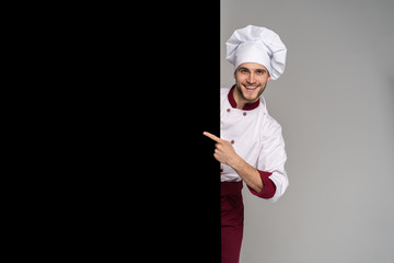 Image of smiling young cook in uniform standing isolated over grey wall background. Looking camera pointing.