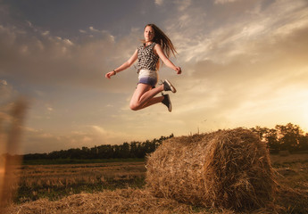 a beautiful, young girl in denim shorts and a plaid shirt jumps off a haystack against the background of the sunset sky and a wheat field