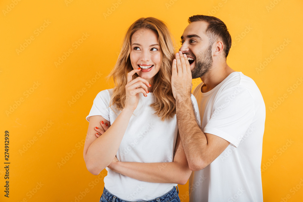 Image of happy couple man and woman in basic t-shirts whispering secrets or gossips to each other - obrazy, fototapety, plakaty 