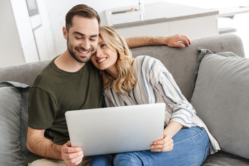 Canvas Print - Happy excited young loving couple sitting on sofa at home using laptop computer.