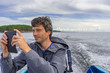 © Tatyana_Andreyeva - Handsome middle-aged man sitting at boat stern, floating along northern lake and using smartphone on beautiful landscape background in summer day. Travel concept. Onega lake, Karelia, Russia