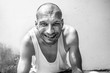 Young positive skinny anorexic bald happy smiling homeless man sitting on the urban street in the city or town near wall with big smile looking at the camera, homelessness social documentary concept