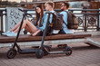 © Fxquadro - Three good happy friends are sitting on the bench at riverside. They have bagpacks and electronic scooters.