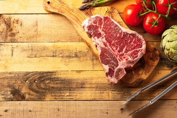 Wall Mural - Fresh raw piece of marbled beef steak with fresh vegetables and tongs, on a cutting board, on a wooden background. Restaurant menu, home menu. Delicious food, gastronomy, culinary background