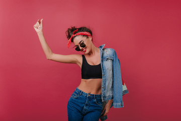 Wall Mural - Enthusiastic sporty girl in sunglasses dancing on claret background. Studio shot of pleased black-haired woman in tank-top and denim jacket.