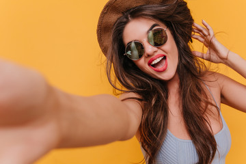 Wall Mural - Close-up portrait of good-looking excited woman taking picture of herself. Funny girl in sunglasses making selfie on yellow background.