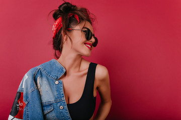 Wall Mural - Beautiful brown-haired girl with black sunglasses laughing on claret background. Studio shot of cute smiling young woman holding denim jacket.