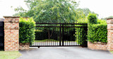 Fototapeta  - Black metal driveway property entrance gates set in brick fence with garden shrubs and trees in background