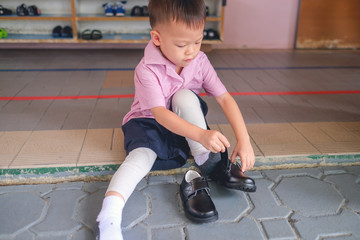 Cute Little Asian 3 - 4 years old kindergarten kid in uniform sitting and concentrate on putting on children's classics boys black leather velcro Shoes, Encourage Self-Help Skills in Children concept