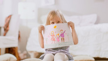 Cute Young Girl Sitting On Pillows And Shows Drawing Of Her Family And Hides Behind It. Sunny Living Room.
