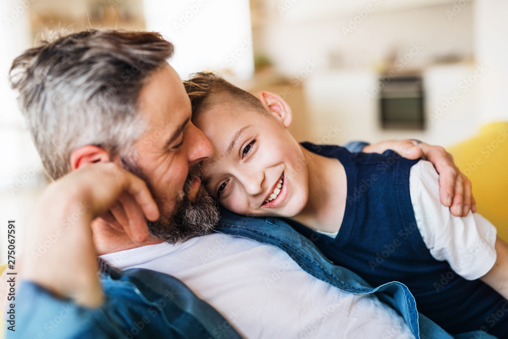 Mature father with small son sitting on sofa indoors, resting. - obrazy, fototapety, plakaty 