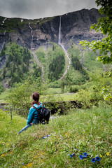 Fotomurales - woman hiker looks at high picturesque waterfalls in lush green forest and mountain landscape