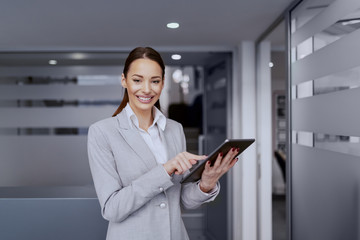 Wall Mural - Charming businesswoman in formal wear and with big toothy smile using tablet while standing in hall at work.