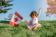 Portrait Of Adorable Cute Little Caucasian Baby Toddler Girl Sitting On Green Grass In Park Outside And Holding Waving Large Canadian Flag. Kid Child Citizen Celebrating Canada Day On 1st Of July.