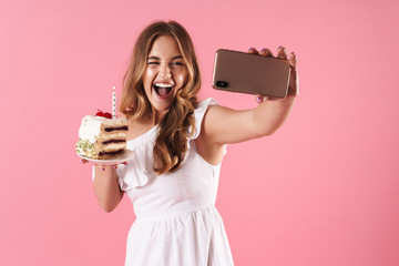 Wall Mural - Image of young laughing woman taking selfie photo on cellphone and winking while holding piece of cake with candle