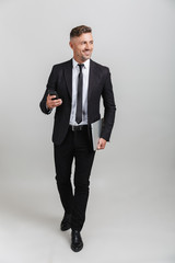 Wall Mural - Full length portrait of happy adult businessman in office suit holding smartphone and laptop