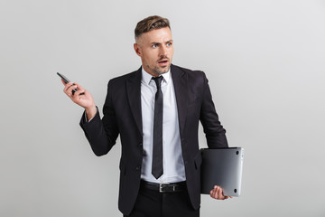 Wall Mural - Portrait of outraged adult businessman in office suit holding smartphone and laptop while standing
