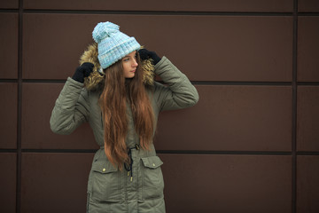 Portrait of a young pretty caucasian teenage girl in a blue hat, black gloves and a green jacket with a hood in the winter outside.