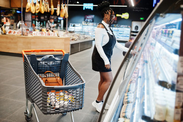 Poster - African woman with shopping cart choose yogurt bottle from fridge at supermarket.