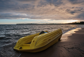 Wall Mural - yellow pedal boat floating ashore and lying upside down on the beach, dark clouds in the sky and beach in the foreground and sunset in the background