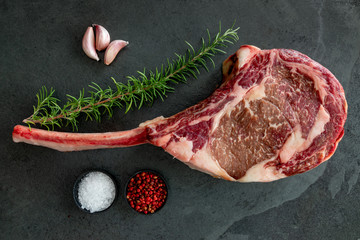 Raw, dry-aged wagyu tomahawk steak on a black stone table with salt, pepper, garlic and rosemary. Top view