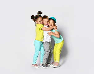 Studio portrait of children on a light background: full body shot of three children in bright clothes, two girls and one boy. Triplets, brother and sisters. hugging on camera. Family ties, friendship