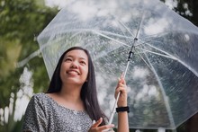 Asian Beautifil Smiling Woman Covering Umbrellas In The Rain With Green Tree Park Background.Concept Of Preparing For The Rainy Season.