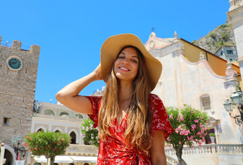 Wall Mural - Summer holiday in Italy. Portrait of young woman with straw hat and red dress with Taormina village on the background, Sicily, Italy.