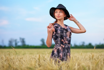 Wall Mural - Cute attractive young girl with black hat on wheat field during sunset. Pensive look. Romantic atmosphere.