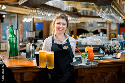 Smiling friendly waitress serving a pint of draft beer in a pub. Portrait of happy young woman serving beer in bar, looking at camera smiling.