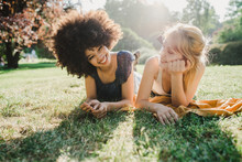 Two Young Women Are Lying On The Grass In A Park While They Are Joking - Back Light Portrait Of Millennials Having A Summer Day At Sunset In The City