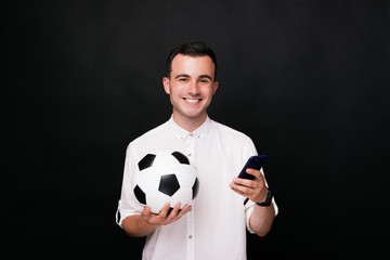 Happy young man smiling at the camera holding his smart phone and a soccer ball on black background. Let's watch the match online on the phone!