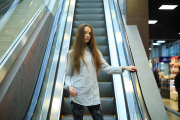 Portrait of a young pretty caucasian teenage girl with long hair in a shirt on the escalator in a mall.