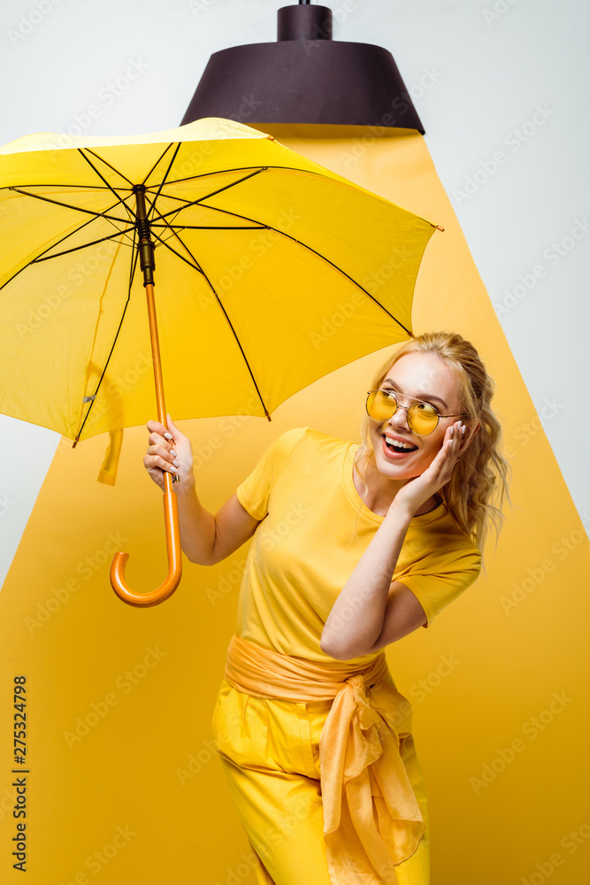 cheerful blonde girl looking at umbrella while touching sunglasses on white and yellow - obrazy, fototapety, plakaty 