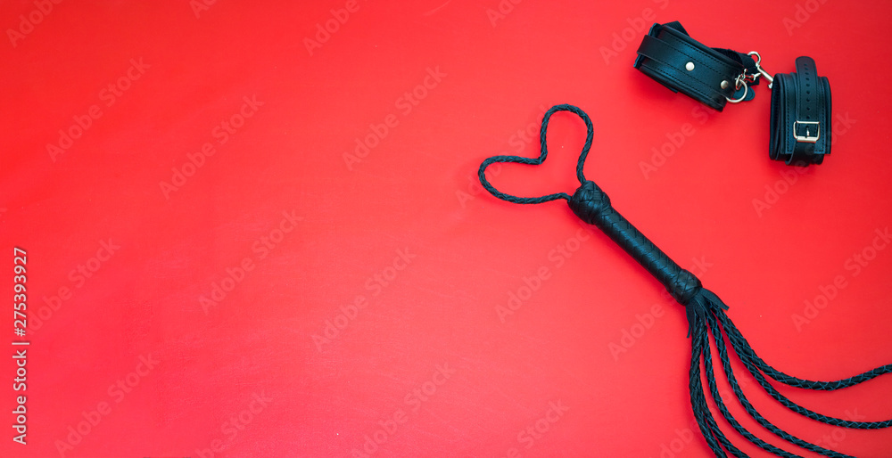 Accessories for bdsm on a red leather background. Lash in the shape of a heart and leather handcuffs. Valentine's Day. Erotic shop. Copy space - obrazy, fototapety, plakaty 