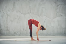 Side View Of Beautiful Caucasian Brunette In Red Sports Wear Standing Barefoot On The Mat In Standing Forward Bend Yoga Posture.