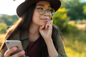 Poster - Photo of attractive woman wearing stylish hat and eyeglasses using cellphone while walking in green park