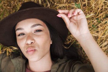 Poster - Photo from top of charming woman wearing elegant hat making kissing lips at camera while lying on grass outdoors