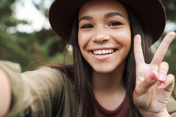 Poster - Photo closeup of happy woman wearing hat smiling and showing peace sign while taking selfie photo outdoors