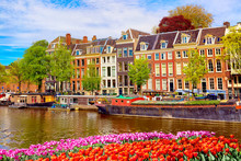 Cityscape View Of The Canal Of Amsterdam In Summer With A Blue Sky And Traditional Old Houses. Colorful Spring Tulips Flowerbed On The Foreground. Picturesque Of Amsterdam, The Netherlands.