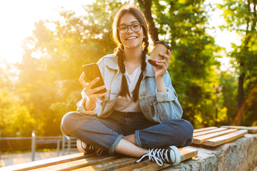 Wall Mural - Happy cute young student girl wearing eyeglasses sitting outdoors in nature park using mobile phone drinking coffee.